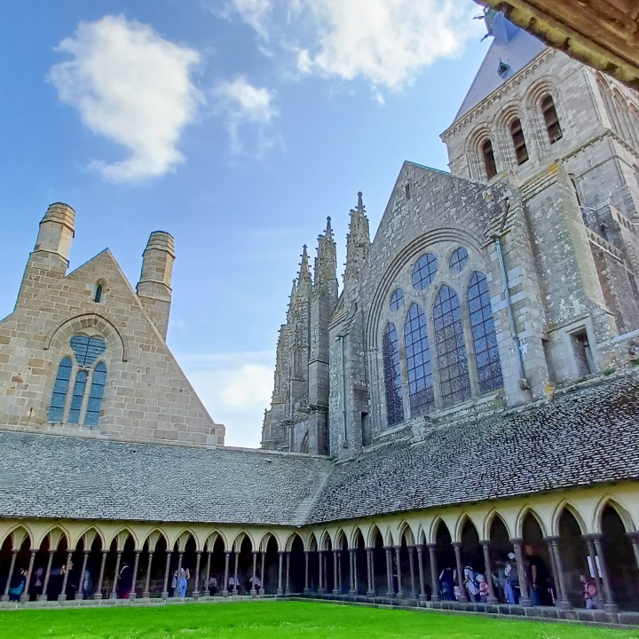 A tranquil view of the cloister at Mont Saint Michel in summer, highlighting its serene Gothic architecture amidst blooming flowers and lush greenery. The historic arches and intricate stonework of the cloister are beautifully complemented by the vibrant summer flora, creating a peaceful atmosphere perfect for contemplation and reflection. This picturesque setting showcases the harmonious blend of natural beauty and medieval craftsmanship that defines Mont Saint Michel.